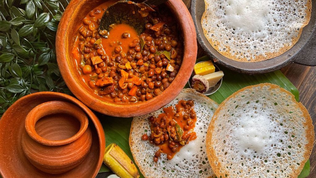 Kerala-style appam with lacy edges served alongside a clay pot of chickpea curry on a banana leaf.