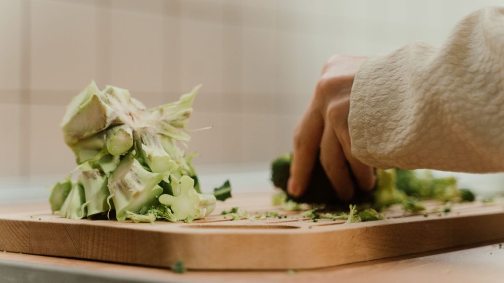 A person chopping broccoli on a wooden board beside a pile of trimmed stalks and leaves.