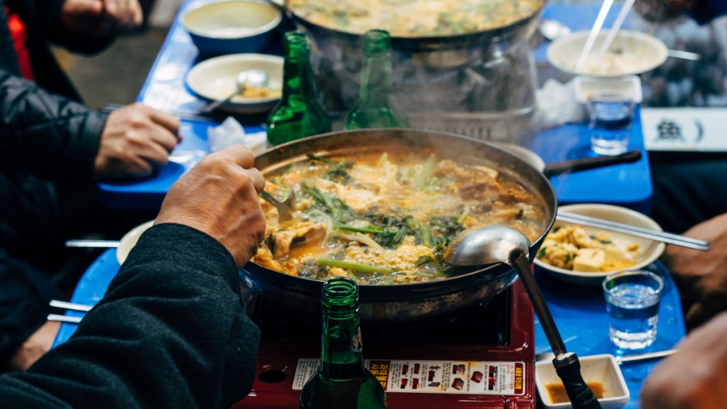Diners sharing a bubbling Korean stew from a wide pan on a tabletop burner, surrounded by side dishes.