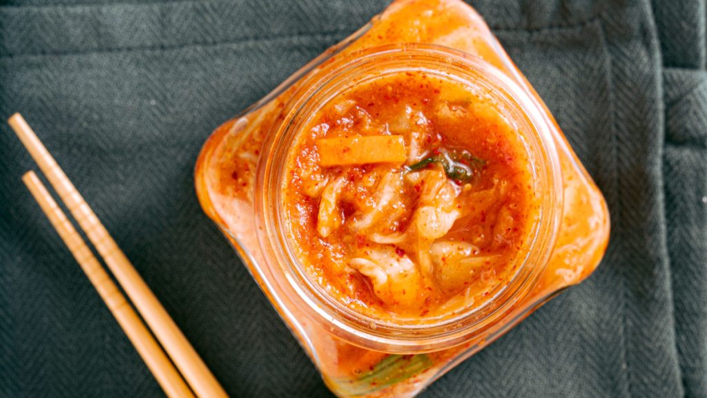 Open glass jar of napa-cabbage kimchi beside wooden chopsticks, photographed from above.
