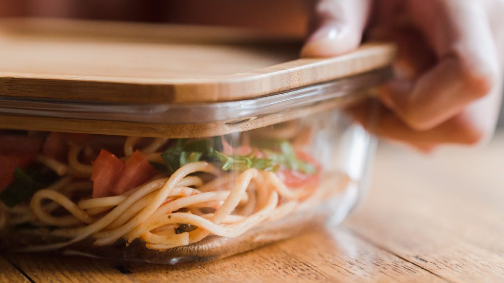 A hand placing a wooden lid on a glass container holding leftover noodles, tomatoes and greens.