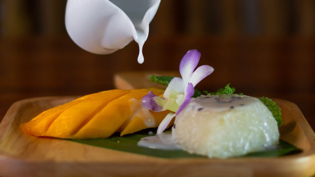 Sliced ripe mango beside Thai sticky rice as coconut cream is poured over it on a banana leaf-lined wooden plate.