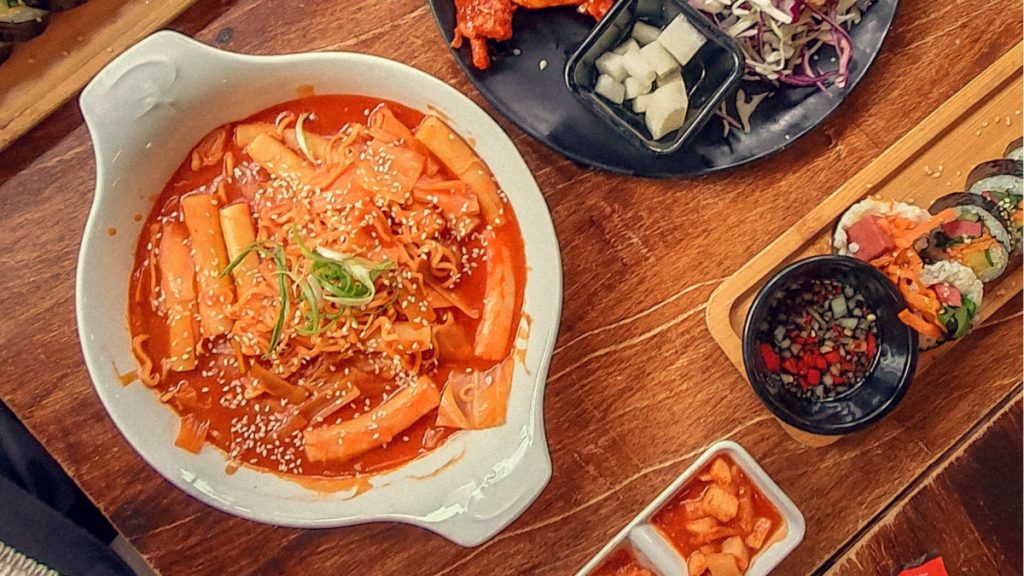Tteokbokki rice cakes in red sauce with sesame and spring onion, served beside kimbap and kimchi.
