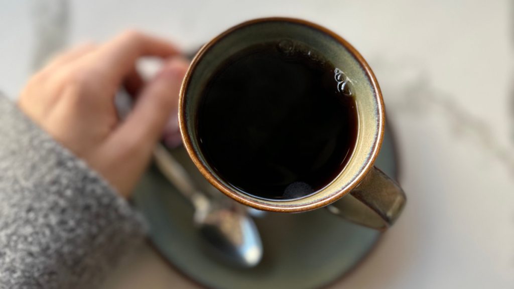 Person sitting beside a ceramic mug of black coffee on a matching saucer, photographed from above.