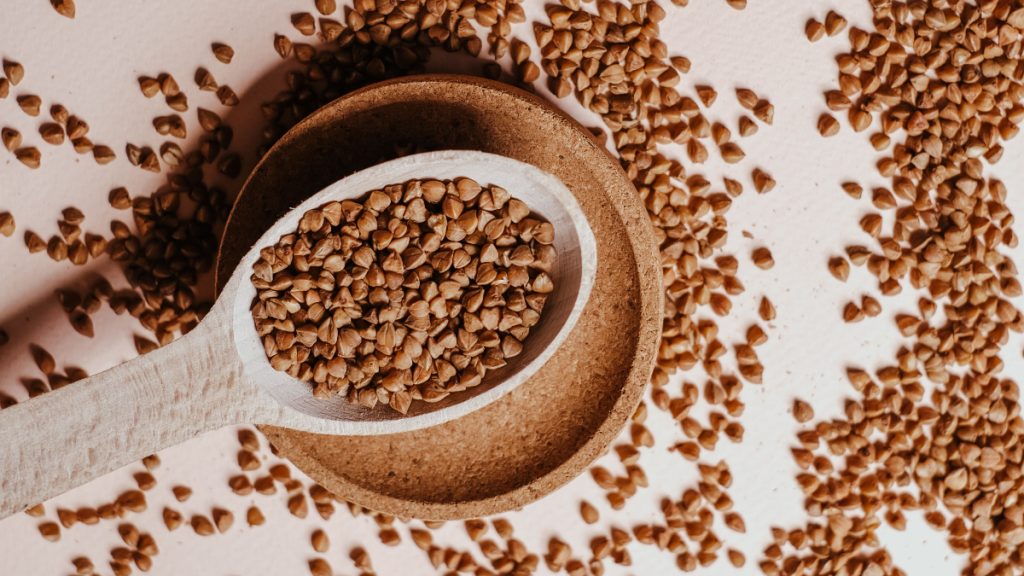 A spoonful of buckwheat is lifted from a shallow dish containing more grains.