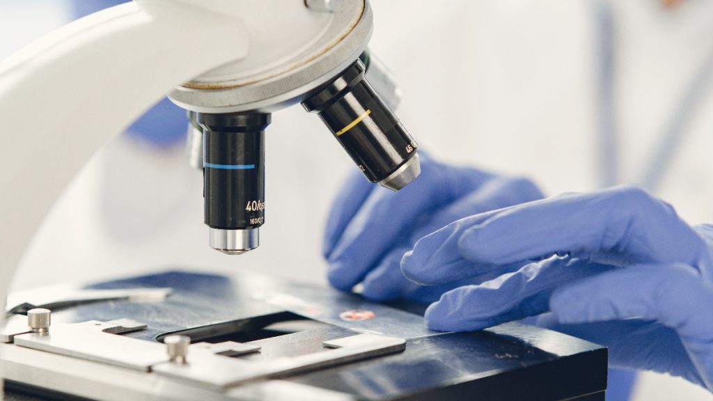 Close-up of a microscope’s objective lenses and stage beside blue-gloved hands.