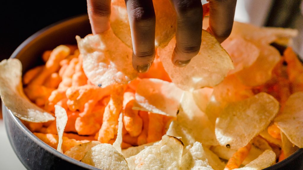 A hand reaching into a bowl of crisps and orange puffed snacks.