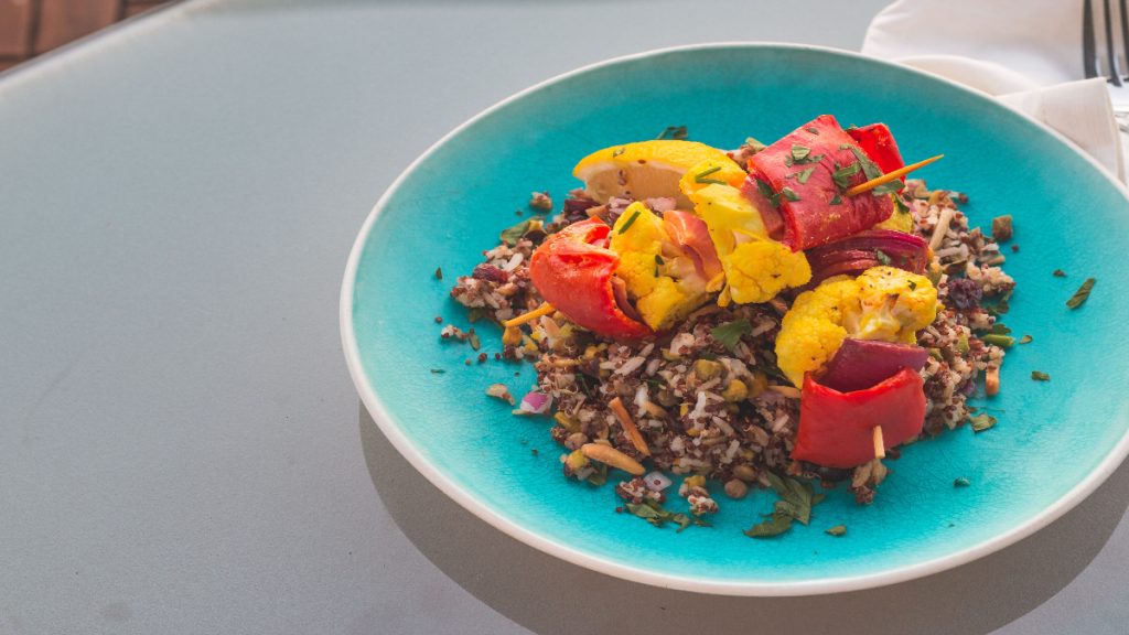 Mixed-grain salad topped with skewers of roasted cauliflower and red and yellow peppers on a turquoise plate.